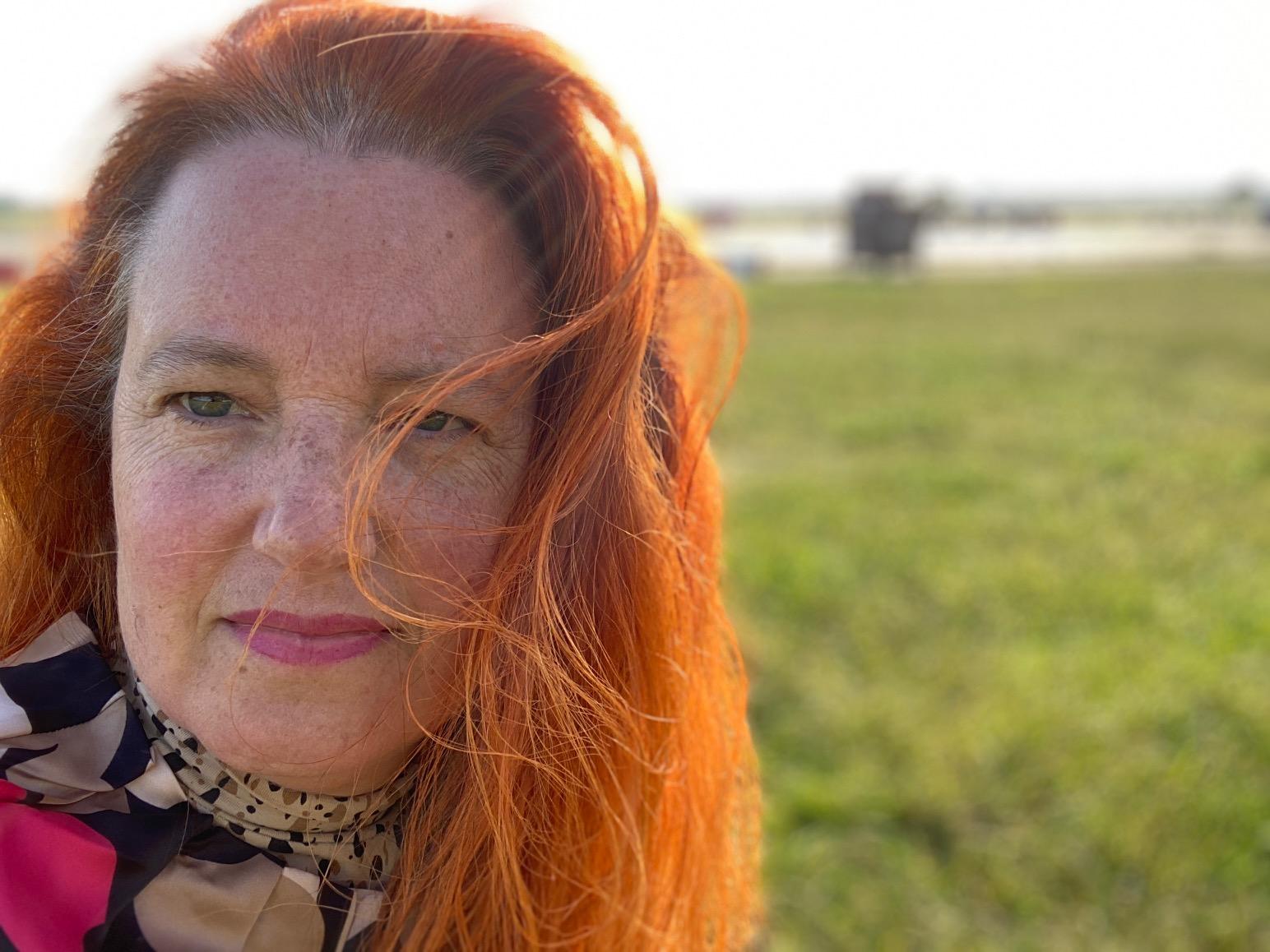 Maria Brasse mit roten Haaren und buntem Oberteil. Im Hintergrund unscharf grüne Wieder und Himmel